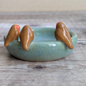 K&ouml;nnte beinhalten: Eine runde, hellblaue Keramikschale mit gesprenkelter Oberfl&auml;che, auf deren Rand drei kleine, braune, geformte V&ouml;gel sitzen. Die V&ouml;gel haben glatte, gl&auml;nzende Oberfl&auml;chen und ein Vogel zeigt einen kleinen, leuchtend orangen Fleck am Kopf.
