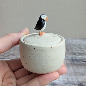 May include: A small, round, off-white ceramic jar with a speckled finish and a matching lid. A hand-painted, three-dimensional puffin figurine with black, white, and orange details is attached to the centre of the lid as a decorative handle.