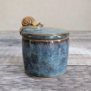 May include: A ceramic jar with a lid, glazed in shades of blue and brown. A detailed snail figurine sits atop the lid. The jar has a rounded shape and a textured surface, suitable for storing small items.