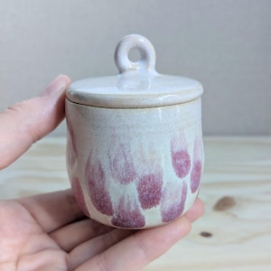 May include: A small, cylindrical ceramic jar with a matching lid featuring a circular handle. The jar has a cream-colored base glaze decorated with scattered, vertical, brush-like strokes in a muted, dusty rose pink color throughout the exterior.