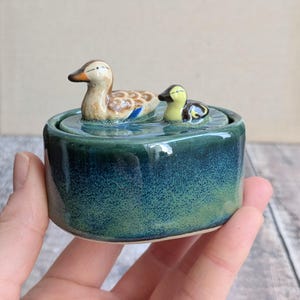 May include: A ceramic trinket box with a teal and green glaze. The lid features two duck figurines, one adult and one duckling, floating on a water-like surface. The ducks have brown, yellow, and blue details.
