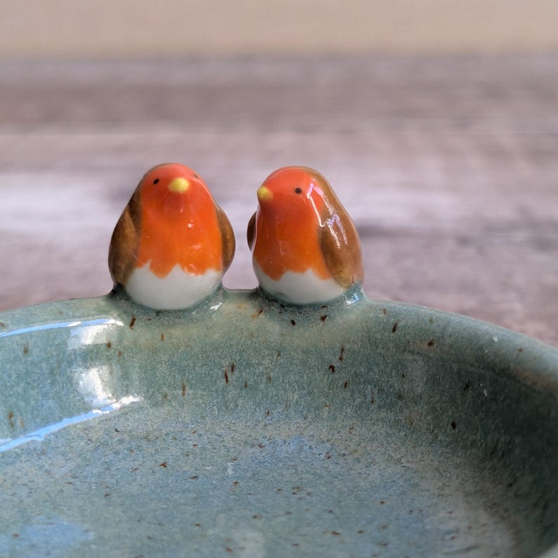 K&ouml;nnte beinhalten: Eine Keramikschale mit zwei kleinen, handbemalten Rotkehlchen-Figuren am Rand. Die Schale hat eine gesprenkelte, hellblau-gr&uuml;ne Glasur, w&auml;hrend die V&ouml;gel leuchtend orangefarbene Brustpartien, braune Fl&uuml;gel, wei&szlig;e B&auml;uche und gelbe Schn&auml;bel haben.