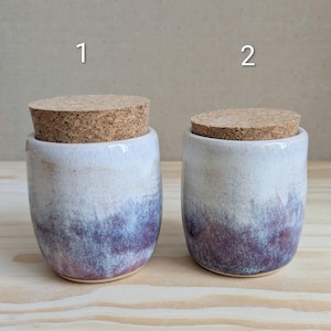 May include: Two ceramic jars with cork lids, labelled 1 and 2. Each jar features a cylindrical shape with a white upper section that transitions into a mottled purple and blue base. The surface has a smooth, glazed finish with visible colour gradients.