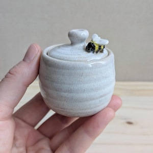 May include: A small, round, off-white ceramic jar with a matching lid. The jar features a subtle horizontal ribbed texture. A tiny, hand-sculpted bumblebee with black and yellow stripes and white wings is attached to the top of the lid.