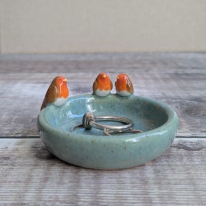 K&ouml;nnte beinhalten: Eine runde, gesprenkelte hellblaue Keramikschale als Ablage, verziert mit drei kleinen, geformten Rotkehlchen-Figuren am Rand. Die V&ouml;gel haben orangefarbene Brustpartien, braune Fl&uuml;gel und wei&szlig;e B&auml;uche. Zwei silberne Ringe liegen in der Schale.