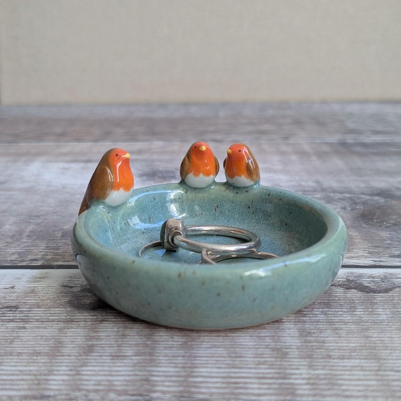 K&ouml;nnte beinhalten: Eine runde, gesprenkelte hellblaue Keramikschale als Ablage, verziert mit drei kleinen, geformten Rotkehlchen-Figuren am Rand. Die V&ouml;gel haben orangefarbene Brustpartien, braune Fl&uuml;gel und wei&szlig;e B&auml;uche. Zwei silberne Ringe liegen in der Schale.