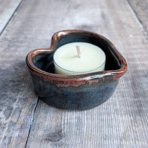 May include: A heart-shaped ceramic candle holder with a dark blue glaze and a reddish-brown rim. A white tea light candle sits inside the holder. The candle holder is on a wooden surface.