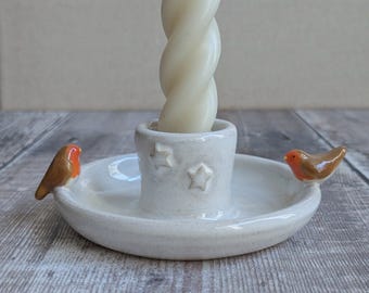 May include: A white ceramic candle holder featuring a central socket decorated with embossed stars. Two small, brown and orange robin figurines are attached to opposite sides of the circular base. A single, off-white twisted taper candle is inserted.