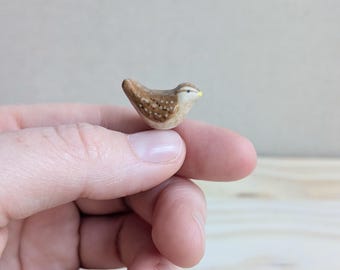 Könnte beinhalten: Eine Miniatur-Vogelfigur aus Keramik mit braunem Körper, weißen Sprenkeln, einer hellbeigen Brust und einem kleinen gelben Schnabel. Der Vogel ist in einer ruhenden Position geformt und weist eine glatte, glänzende Oberfläche auf.