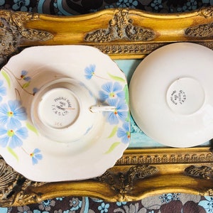 K&ouml;nnte beinhalten: Ein Vintage-Teetassen- und Untertassen-Set mit einem Blumendesign mit blauen und wei&szlig;en Blumen. Die Untertasse hat einen gewellten Rand. Die Teetasse tr&auml;gt auf der Unterseite die Worte "Phoenix Ware Made in England T.T. & S. Ltd.".
