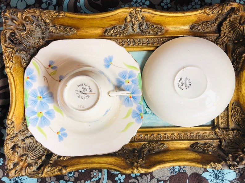 K&ouml;nnte beinhalten: Ein Vintage-Teetassen- und Untertassen-Set mit einem Blumendesign mit blauen und wei&szlig;en Blumen. Die Untertasse hat einen gewellten Rand. Die Teetasse tr&auml;gt auf der Unterseite die Worte "Phoenix Ware Made in England T.T. & S. Ltd.".