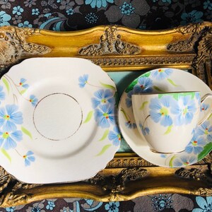 K&ouml;nnte beinhalten: Ein Vintage-Teetassen- und Untertassen-Set mit floralem Design. Tasse und Untertasse sind wei&szlig; mit blauen und gr&uuml;nen Blumen. Das Set wird auf einem goldfarbenen Tablett pr&auml;sentiert.