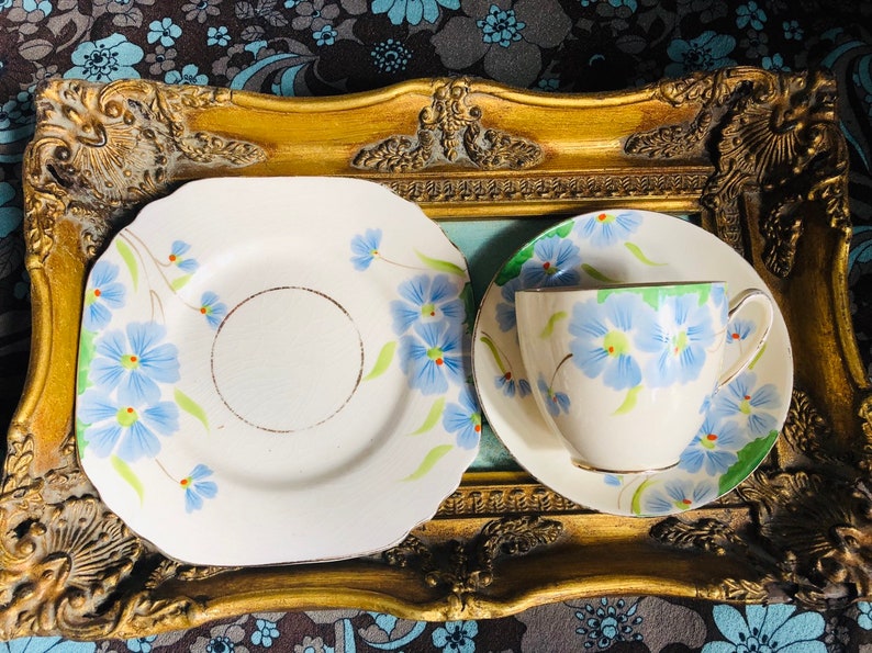 K&ouml;nnte beinhalten: Ein Vintage-Teetassen- und Untertassen-Set mit floralem Design. Tasse und Untertasse sind wei&szlig; mit blauen und gr&uuml;nen Blumen. Das Set wird auf einem goldfarbenen Tablett pr&auml;sentiert.