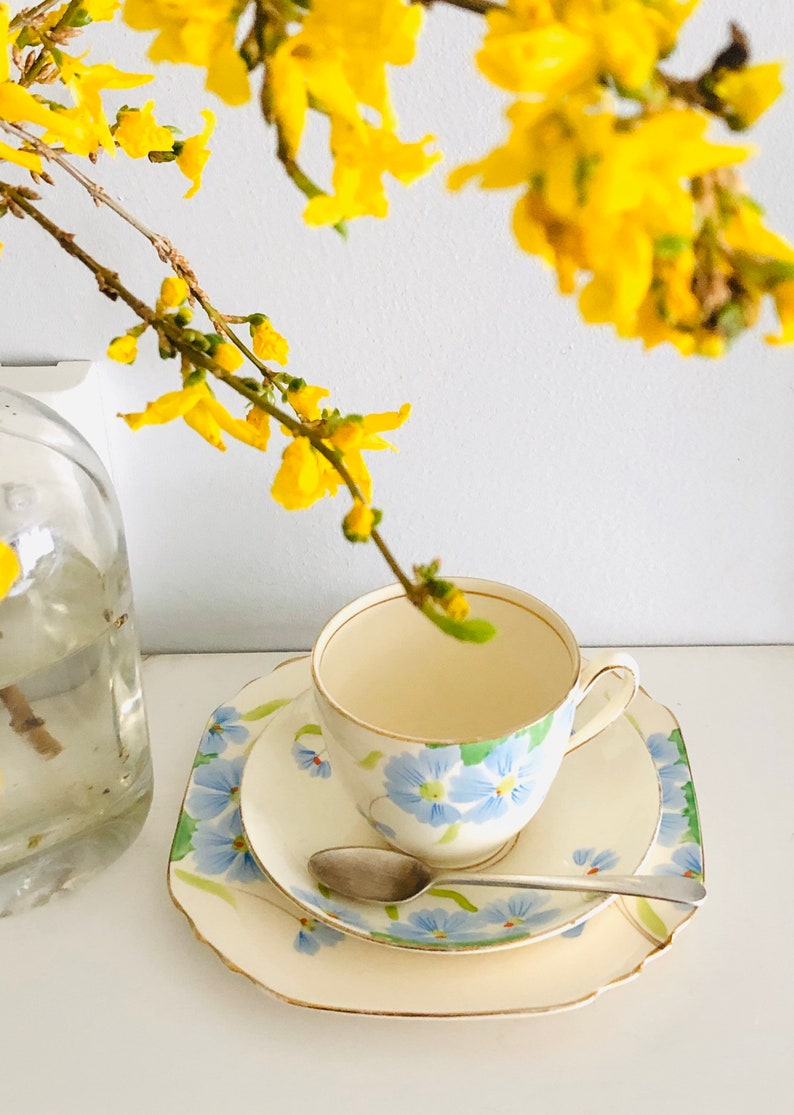 K&ouml;nnte beinhalten: Ein wei&szlig;es Tee-Set mit einem blauen Blumenmuster. Das Set befindet sich auf einer wei&szlig;en Oberfl&auml;che mit einem Zweig gelber Blumen.