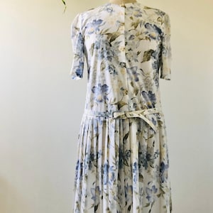Può includere: Abito vintage con stampa floreale su manichino. L'abito presenta uno sfondo bianco con motivi floreali blu e verdi. Ha maniche corte, un'abbottonatura frontale e una gonna a pieghe. Una cintura abbinata segna la vita.