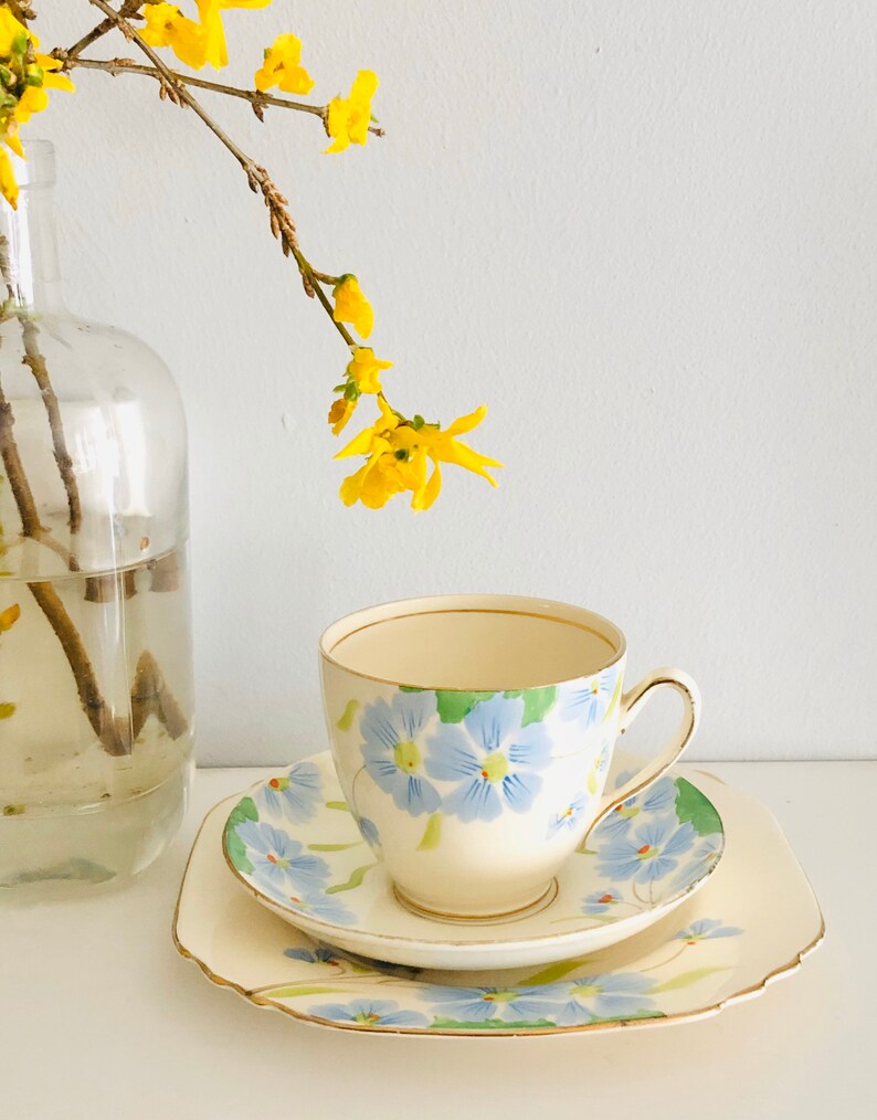 K&ouml;nnte beinhalten: Ein Vintage-Teetassen- und Untertassen-Set mit Blumenmuster. Tasse und Untertasse sind wei&szlig; mit blauen und gr&uuml;nen Blumen. Die Untertasse hat einen gewellten Rand.