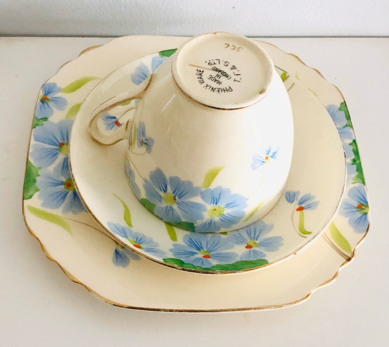 K&ouml;nnte beinhalten: Ein dreiteiliges Teeset mit Blumenmuster. Das Set besteht aus einer Tasse, einer Untertasse und einem gr&ouml;&szlig;eren Teller. Tasse und Untertasse haben ein blaues und gr&uuml;nes Blumenmuster mit goldener Verzierung. Der gr&ouml;&szlig;ere Teller hat ein &auml;hnliches Muster, ist aber gr&ouml;&szlig;er. Auf der R&uuml;ckseite der Tasse steht "PHOENIX WARE MADE IN ENGLAND T.T. & S. LTD. 322".