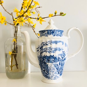 May include: A blue and white ceramic teapot with a floral design and a scene of a village. The teapot has a handle and a spout. The teapot is sitting on a white surface.