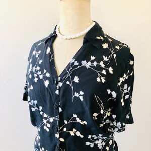 May include: A black button-up shirt with a floral print featuring white and light blue flowers. The shirt has short sleeves and a collared neckline. A white pearl necklace is worn on a mannequin wearing the shirt.