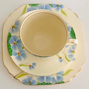 K&ouml;nnte beinhalten: Ein Vintage-Teetassen- und Untertassen-Set mit floralem Design. Tasse und Untertasse sind wei&szlig; mit blauen und gr&uuml;nen Blumen. Die Untertasse hat einen gewellten Rand.