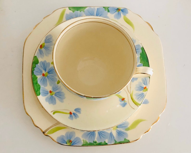 K&ouml;nnte beinhalten: Ein Vintage-Teetassen- und Untertassen-Set mit floralem Design. Tasse und Untertasse sind wei&szlig; mit blauen und gr&uuml;nen Blumen. Die Untertasse hat einen gewellten Rand.