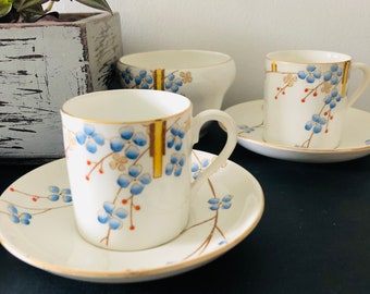 Könnte beinhalten: Ein Set aus drei weißen Porzellan-Teetassen und Untertassen mit einem zarten Blumenmuster in Blau-, Rot- und Goldtönen. Die Tassen und Untertassen haben einen goldenen Rand.