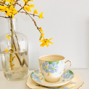 K&ouml;nnte beinhalten: Ein wei&szlig;es Tee-Set mit Untertasse mit einem blauen Blumenmuster. Tasse und Untertasse haben einen goldenen Rand. Im Hintergrund steht eine Vase mit gelben Blumen.