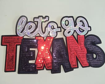May include: Let's go Texans! Red, black and white sequined letters on a black background.