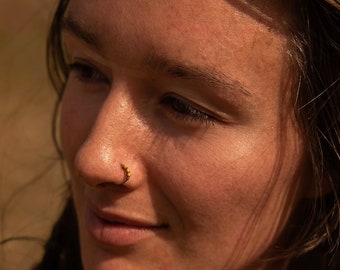 May include: A woman with long brown hair styled in dreadlocks, wearing a brown sweater, looks off to the side with a slight smile. She has a small gold nose ring.