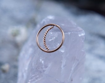 May include: A gold-toned nose ring with a twisted wire design. The ring is displayed on a white crystal.