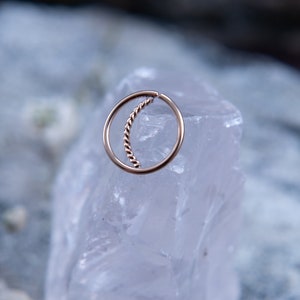 May include: A gold-toned nose ring with a twisted wire design. The ring is displayed on a white crystal.