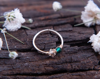 May include: A silver ring with a clear crystal and a green gemstone. The ring is on a rustic wooden surface with white flowers.