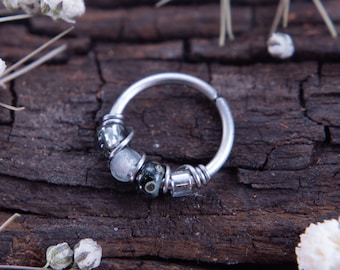 May include: A silver hoop earring with a beaded design featuring black, white, and gray stones. The earring is on a wooden surface with white flowers.
