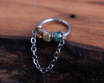 May include: A silver nose ring with a chain and three colorful beads. The beads are yellow, green, and blue.