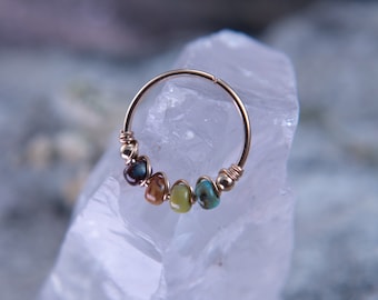 May include: A gold hoop nose ring with five colorful gemstone beads. The beads are a dark blue, brown, yellow, green, and turquoise.
