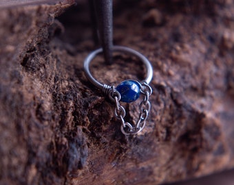 May include: A silver ring with a blue gemstone and a dangling chain. The ring is being held by a pair of tweezers.