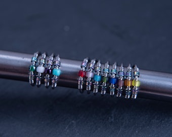 May include: A set of 16 silver rings with colorful glass beads. The rings are stacked on a metal surface. The beads are in various colors, including red, blue, green, yellow, and purple.