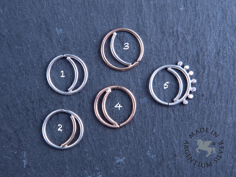 May include: Five silver and rose gold half-moon septum rings with numbers 1 through 5. The rings are on a dark gray background. Made in Argentium Silver.