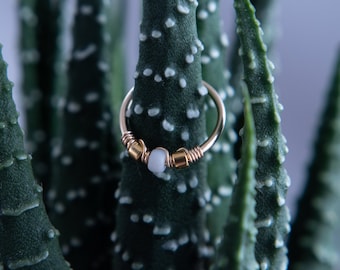 May include: A gold hoop earring with a white stone and gold wire accents. The earring is shown against a green plant background.