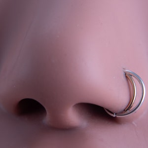 May include: A close-up of a silver nose ring with a double hoop design. The ring is worn in a person's nose.