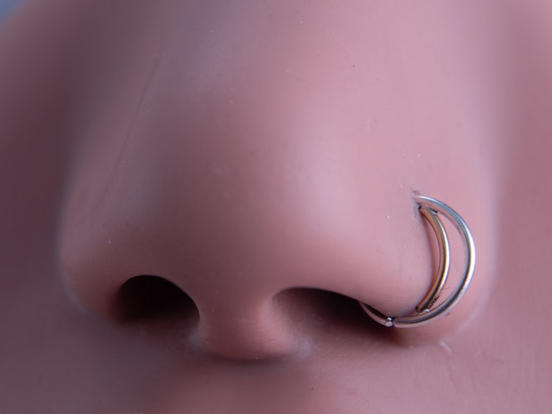 May include: A close-up of a silver nose ring with a double hoop design. The ring is worn in a person's nose.