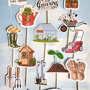 May include: A selection of garden-themed cake toppers on wooden sticks. The toppers feature a watering can with a frog, a greenhouse, a wheelbarrow, a lawnmower, and the text "It's Time for Gardening."