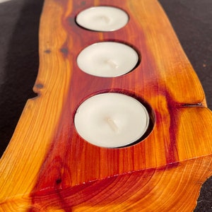 May include: A handcrafted wooden candle holder with four circular indentations, each holding a white tea light candle. The holder is made from a piece of natural wood, showcasing a warm orange and red grain pattern. The edges are roughly cut, giving it a rustic appearance.