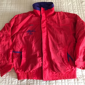 May include: A red and purple windbreaker jacket with a zipper closure and a drawstring at the waist. The jacket has a logo on the front that says "Mountain Tops" and "Skiing Life".