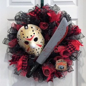 May include: A decorative wreath featuring a hockey mask with red markings, a grey machete with red blood splatters, and black and red mesh ribbons. A small rectangular sign reads Welcome to Camp Crystal Lake Est 1935.