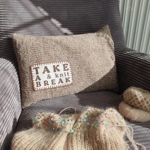 Könnte beinhalten: Ein beiges Strickkissen mit dem Text "TAKE A & knit BREAK" auf einem weißen Aufnäher. Das Kissen liegt auf einem grauen Cord-Sessel, mit Garn und einem Strickkleidungsstück mit Pailletten.