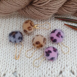 Könnte beinhalten: Fünf dekorative Maschenmarkierer mit Kunstfell-Pompons in verschiedenen Farben, darunter Beige, Lila und Rosa, jeweils an einer goldfarbenen Sicherheitsnadel befestigt. Die Markierer sind auf einer weißen Strickoberfläche angeordnet, mit einem Wollknäuel im Hintergrund.
