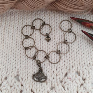 Könnte beinhalten: Ein bronzefarbenes Armband mit kreisförmigen Gliedern und dekorativen Anhängern. Zu den Anhängern gehören ein kleines Herz und eine Lotusblume. Das Armband wird auf einer cremefarbenen, strukturierten Oberfläche mit Garn und Stricknadeln im Hintergrund präsentiert.