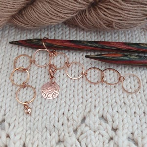 Könnte beinhalten: Eine Sammlung von Maschenmarkierern aus Roségold und ein Charm mit Herz und kreisförmigem Design. Zwei Stricknadeln aus Holz sind ebenfalls im Bild zu sehen. Die Artikel sind auf einer cremefarbenen Strickoberfläche angeordnet, mit einem Knäuel Wolle im Hintergrund.