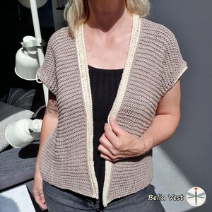 Puede incluir: Un chaleco de crochet beige de manga corta con ribete color crema. El chaleco está abierto por delante y tiene un diseño texturizado y acanalado. El texto "Belia Vest" es visible en la esquina inferior derecha.